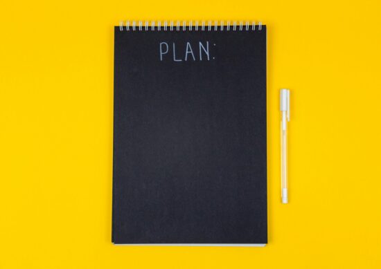 Wide view image of blank black spiral note pad and white marker with calligraphic inscription plan on yellow background